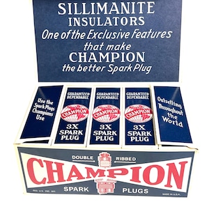 May include: An open box of Champion spark plugs. The box is navy blue and white with red lettering. The box reads "Champion Spark Plugs" and "Sillimanite Insulators". The box contains multiple individual spark plug boxes.