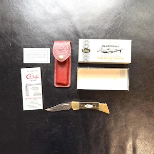 Könnte beinhalten: Ein Case Hammerhead Klappmesser mit braunem Griff und Messingakzenten, präsentiert mit einer roten Lederscheide und Originalverpackung. Die Messerklinge hat ein detailliertes Design.