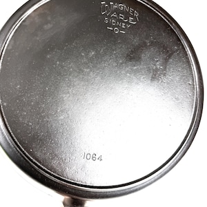 May include: A black cast iron Wagner Ware Sidney-O skillet, with the number 1064 stamped on the base. The skillet has a round cooking surface and a handle.
