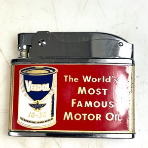 Op de afbeelding: Een zilveren aansteker met een rood en gouden ontwerp. De aansteker heeft een vintage oliedoos met de tekst "Veedol" en "The World's Most Famous Motor Oil".