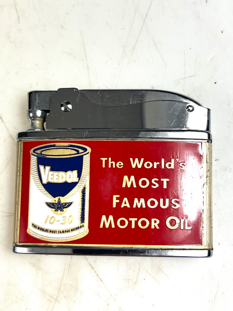 Op de afbeelding: Een zilveren aansteker met een rood en gouden ontwerp. De aansteker heeft een vintage oliedoos met de tekst "Veedol" en "The World's Most Famous Motor Oil".