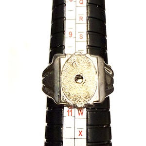 May include: A silver-toned ring with a textured, oval-shaped center stone is placed on a black ring sizer. The ring is positioned at size 11 on the sizer. The ring has a rectangular design with rounded edges.