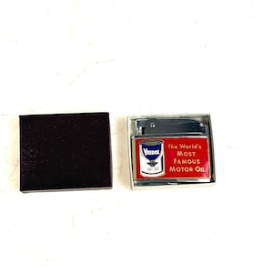 Op de afbeelding: Een zilveren aansteker met een rood en wit Veedol-motoroliedoosontwerp. De tekst op de aansteker luidt "The World's Most Famous Motor Oil".