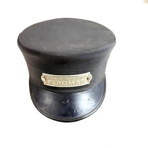 Op de afbeelding: Een zwarte Lackawanna Railroad-vlaggenman's hoed. De hoed heeft een afgeronde bovenkant, een korte rand en een band met een metalen naamplaatje met de tekst "Lackawanna Railroad FLAGMAN."