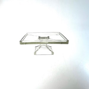 Könnte beinhalten: Ein transparenter Kuchenständer aus Glas mit quadratischer Oberseite und einem dekorativen Sockel. Der Ständer ist für das Präsentieren von Kuchen oder anderen Desserts gedacht.
