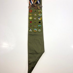 May include: A green sash with a variety of merit badges and awards. The sash is folded in half and the badges are arranged in rows. The badges include a variety of subjects, such as camping, first aid, and citizenship.