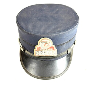 Op de afbeelding: Een marineblauwe conducteurshoed met een zwarte rand en een metalen badge. De badge heeft de tekst "CONDUCTOR" en het nummer "82546". De hoed is een vintage item.