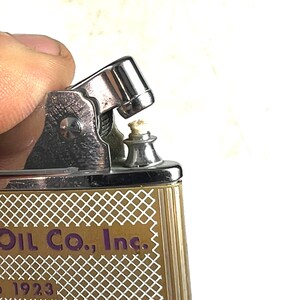 Op de afbeelding: Een vintage zilveren aansteker met een gouden en wit patroon. De aansteker heeft de tekst "Oil Co., Inc." en "to serve you" erop gedrukt.