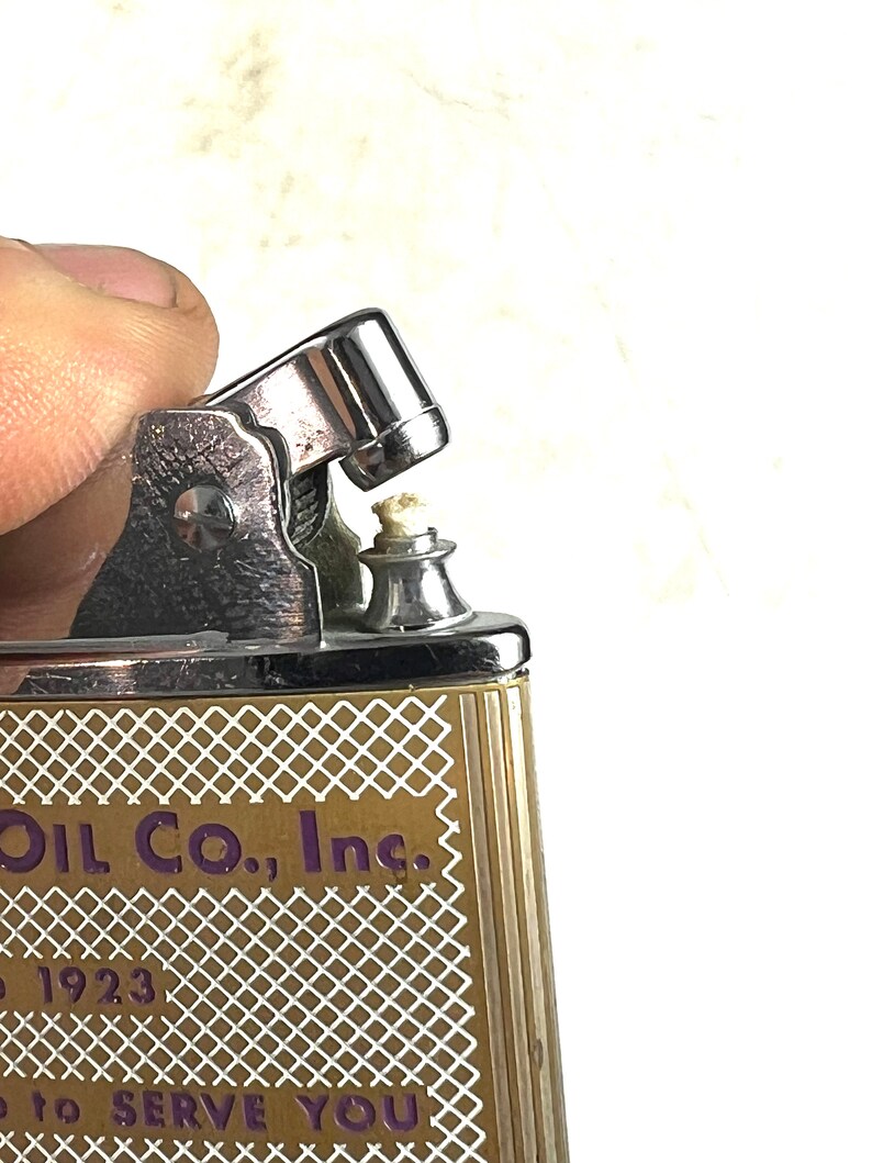 Op de afbeelding: Een vintage zilveren aansteker met een gouden en wit patroon. De aansteker heeft de tekst "Oil Co., Inc." en "to serve you" erop gedrukt.