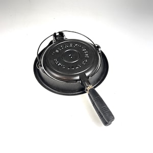 Może przedstawiać: Czarny waflownik z żeliwa z drewnianym uchwytem. Waflownik ma okrągły kształt z napisem "Sidney's 1931 Cast Iron" wokół krawędzi.