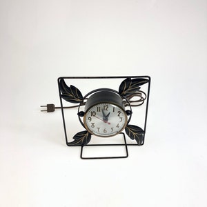 May include: A black metal clock stand with gold leaf accents and a round clock face with black hands and white numbers. The clock is plugged in with a cord.