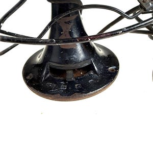 May include: Close-up of a vintage black electric fan. The fan has a circular base, a cylindrical body, and curved blades. The fan's metal components show signs of age and wear, with some rust visible. The fan's design is a classic example of early 20th-century technology.