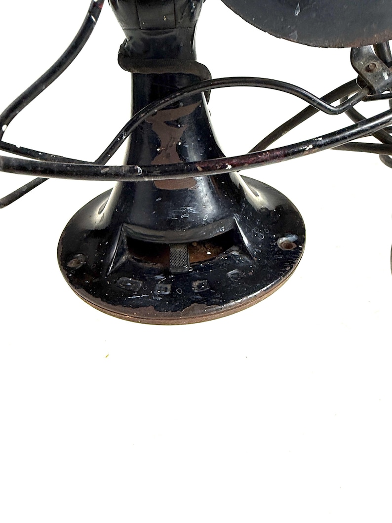 May include: Close-up of a vintage black electric fan. The fan has a circular base, a cylindrical body, and curved blades. The fan's metal components show signs of age and wear, with some rust visible. The fan's design is a classic example of early 20th-century technology.