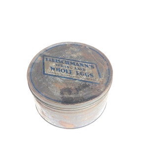 May include: A vintage, rusty, metal tin container with a blue and white label that reads "Fleischmann's Spring Laid Whole Eggs".