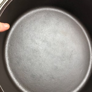 Puede incluir: Una olla de hierro fundido negra con un interior liso y oscuro. La olla est&aacute; vac&iacute;a y lista para cocinar.