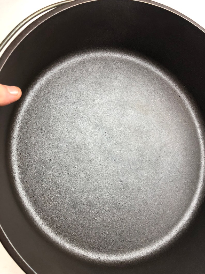 Puede incluir: Una olla de hierro fundido negra con un interior liso y oscuro. La olla est&aacute; vac&iacute;a y lista para cocinar.