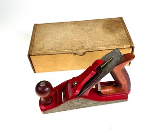 Könnte beinhalten: Ein roter Holzhobel mit einem Holzgriff und einer Metallklinge. Der Hobel befindet sich in seiner Originalverpackung.