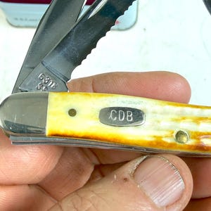 May include: A close-up of a pocket knife with a partially serrated blade. The handle is a cream color with a brown gradient. The knife has the initials "CDB" on a small metal plate.