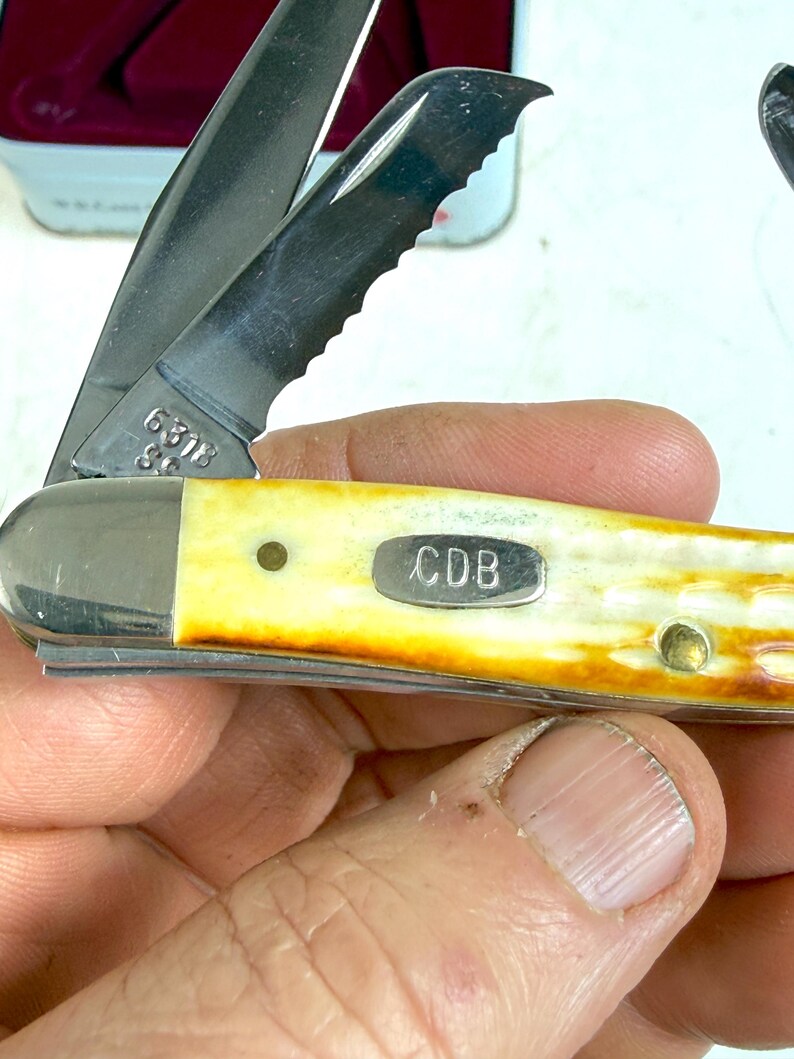 May include: A close-up of a pocket knife with a partially serrated blade. The handle is a cream color with a brown gradient. The knife has the initials "CDB" on a small metal plate.