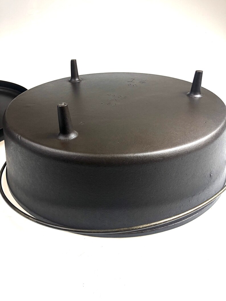Puede incluir: Cazuela de hierro fundido negro con tres patas y un asa de alambre. El fondo de la olla est&aacute; marcado con el texto "14" y "MADE IN USA".