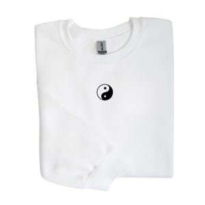 Może przedstawiać: Złożona biała bluza z okrągłym dekoltem z czarno-białym symbolem yin-yang na piersi. Widoczny jest kołnierz i metka z marką. Odzież jest starannie złożona.