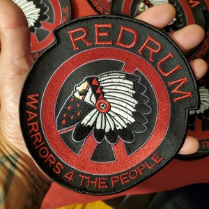 Könnte beinhalten: Schwarz-roter, gestickter Aufnäher mit dem Wort "REDRUM" in Rot, über einem kreisförmigen Design. Das Design beinhaltet einen indianischen Kopfschmuck und den Text "WARRIORS 4 THE PEOPLE".