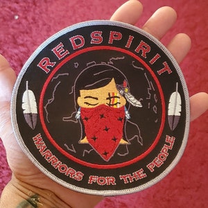 May include: Circular black patch with the words "RED SPIRIT" in red lettering. The patch features a cartoon image of a person wearing a red bandana. The text "WARRIORS FOR THE PEOPLE" is also on the patch. Two white and grey feathers are on the sides.