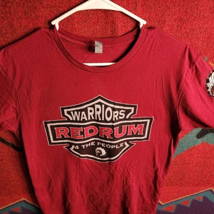 Peut inclure: Un t-shirt bordeaux avec un graphique de logo de style moto qui dit "Warriors Redrum 4 The People". Le logo est en noir, blanc et gris. Il y a deux petites images de coiffures amérindiennes sur les manches.