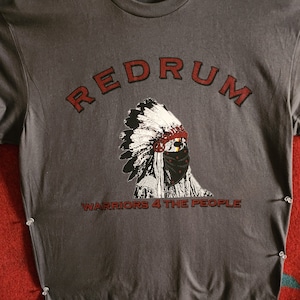 Op de afbeelding: Een grijs T-shirt met een rode afbeelding van een indiaan met een hoofdtooi en een bandana. De tekst "REDRUM" staat boven de afbeelding en "WARRIORS 4 THE PEOPLE" eronder.
