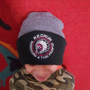 Op de afbeelding: Een grijze en zwarte gebreide beanie met een rood, wit en zwart geborduurd logo met de tekst "Redrum Warriors 4 The People".