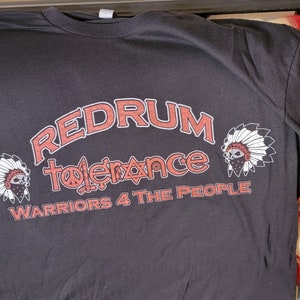 Może przedstawiać: Czarny T-shirt z czerwonym nadrukiem, na którym widnieje napis "Redrum Tolerance Warriors 4 The People". Nadruk ma kształt łuku i zawiera znak pokoju oraz gwiazdę Dawida. Koszulka ma trzy indiańskie nakrycia głowy z przodu i na rękawach.