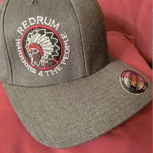 Op de afbeelding: Een zwarte en grijze baseballpet met een rood, wit en zwart geborduurd logo met de tekst "Redrum Warriors 4 the People". Het logo toont een indiaanse hoofdtooi.