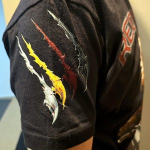 May include: Black t-shirt with a graphic of four stylized claws in silver, red, yellow, and black on the left sleeve. The text "RE" is visible on the right side of the shirt.