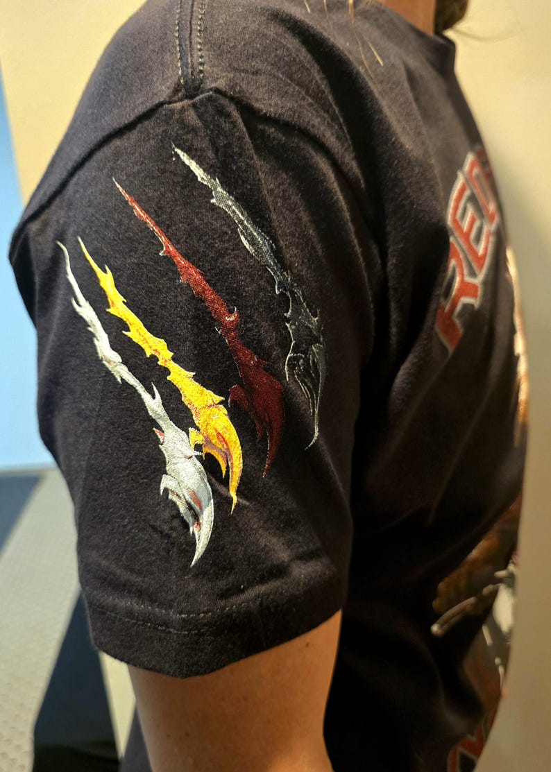 May include: Black t-shirt with a graphic of four stylized claws in silver, red, yellow, and black on the left sleeve. The text "RE" is visible on the right side of the shirt.