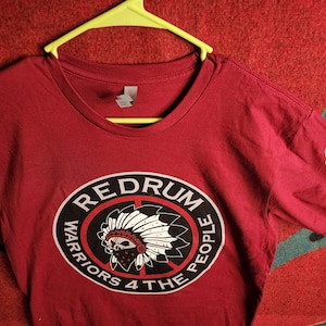 May include: Red t-shirt with a black and white circle graphic featuring a Native American headdress and the text "RED DRUM WARRIORS 4 THE PEOPLE".