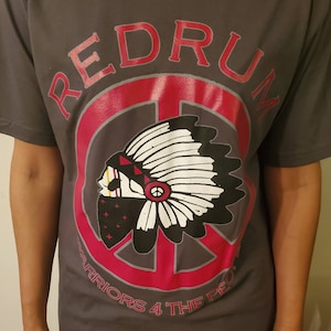Puede incluir: Una camiseta gris con un gráfico de signo de paz rojo y gris. El signo de paz tiene un tocado nativo americano en su interior. El texto "REDRUM" está por encima del signo de paz y el texto "WARRIORS 4 THE PEOPLE" está por debajo del signo de paz.