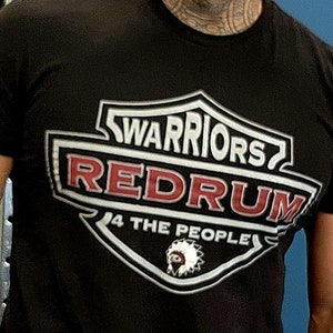 Könnte beinhalten: Schwarzes T-Shirt mit einem weißen und silbernen Schild-Motiv. Der Schild trägt den Text "WARRIORS REDRUM 4 THE PEOPLE" in weißer und roter Schrift. Im Zentrum des Schildes befindet sich ein indianisches Kopfschmuck.
