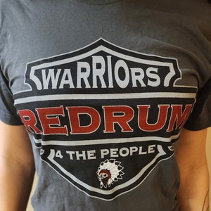Puede incluir: Una camiseta gris con un gráfico en blanco y negro de un escudo con el texto "WARRIORS" y "REDRUM" en rojo. El escudo también tiene el texto "4 THE PEOPLE" y un tocado de nativo americano.
