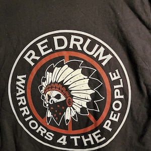May include: Black t-shirt with a white circle graphic. The graphic features a red peace sign with a Native American headdress inside. The text "REDRUM WARRIORS 4 THE PEOPLE" is written around the circle.
