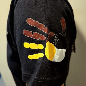 May include: A black t-shirt with a colorful handprint design on the sleeve. The handprint is made up of brown, yellow, and white paint.