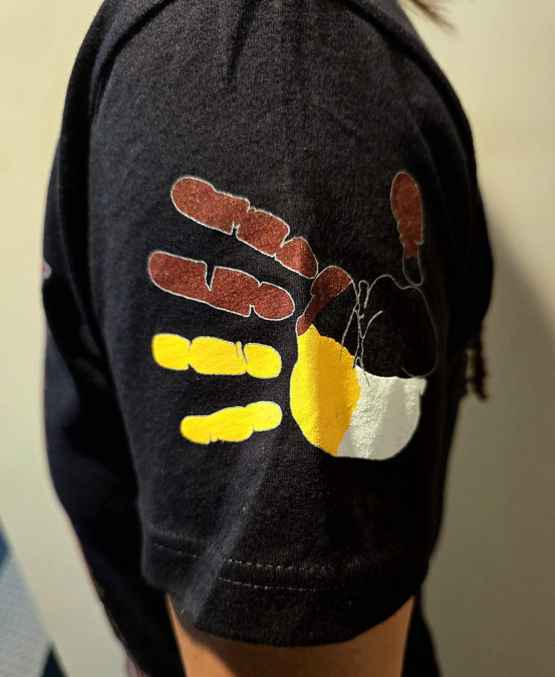 May include: A black t-shirt with a colorful handprint design on the sleeve. The handprint is made up of brown, yellow, and white paint.