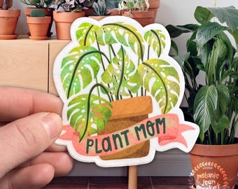 May include: A white sticker with a watercolor illustration of a potted plant with green leaves and the text "Plant Mom" on a pink ribbon.