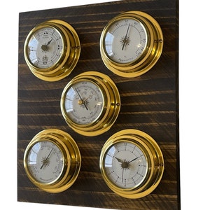 Könnte beinhalten: Ein Satz von fünf Messing-Nautik-Uhren, die auf einer dunkelbraunen Holzplatte montiert sind. Die Uhren haben weiße Zifferblätter mit schwarzen Zeigern und römischen Ziffern. Die Uhren messen Temperatur, Luftfeuchtigkeit, Luftdruck und Zeit.