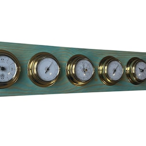 Puede incluir: Una estación meteorológica de pared de madera con cinco instrumentos con bordes de latón. Los instrumentos incluyen un reloj, un barómetro y un termómetro, todos con esferas blancas y agujas negras. La base de madera está pintada de color turquesa.