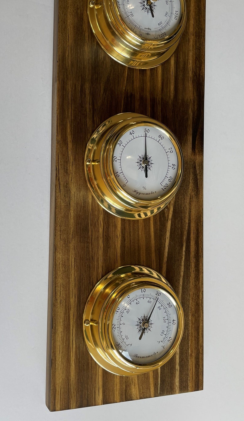 Op de afbeelding: Een messing wandgemonteerde set met een barometer, hygrometer en thermometer. De instrumenten hebben witte wijzerplaten met zwarte markeringen en wijzers. De set is gemonteerd op een donker houten achtergrond.