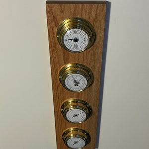 Puede incluir: Un conjunto de cuatro relojes náuticos de latón montados en una placa de madera. Los relojes tienen caras blancas con manecillas negras y marcas de números romanos. Los relojes están dispuestos verticalmente en la placa.