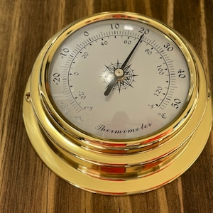 Op de afbeelding: Een messing thermometer met een wit gezicht en zwarte markeringen. De thermometer geeft temperaturen aan van -20 tot 50 graden Fahrenheit en Celsius. De thermometer is gemarkeerd met "Thermometer".