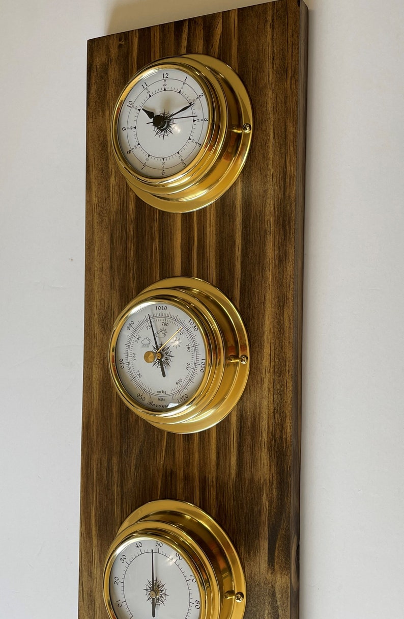 Op de afbeelding: Een set van drie messing nautische klokken gemonteerd op een donkerhouten paneel. De klokken omvatten een barometer, een thermometer en een klok met een kompasroos.