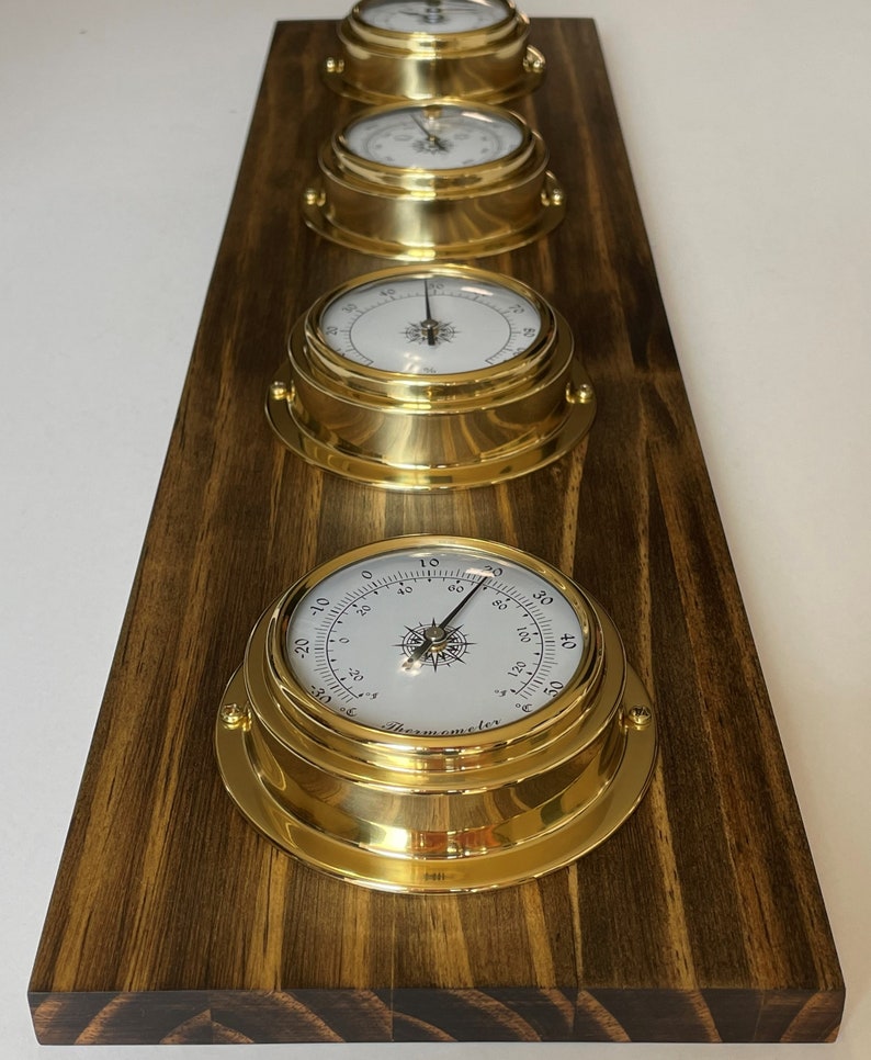 Op de afbeelding: Vier messingkleurige analoge meters gemonteerd op een donkerhouten paneel. De meters hebben witte wijzerplaten met zwarte markeringen en wijzers. De meters meten temperatuur, vochtigheid, luchtdruk en windsnelheid. De meters zijn allemaal in nautische stijl.