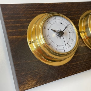 Puede incluir: Dos relojes de barco de lat&oacute;n con caras blancas y manecillas negras montados en una placa de madera marr&oacute;n oscuro.